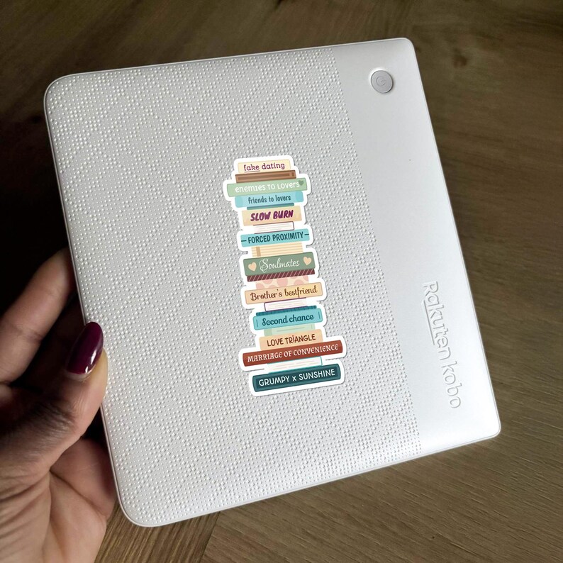 May include: A white Rakuten Kobo e-reader with a stack of colorful stickers. The stickers feature text like "fake dating," "enemies to lovers," and "marriage of convenience." The e-reader is held by a person with a dark red nail polish.