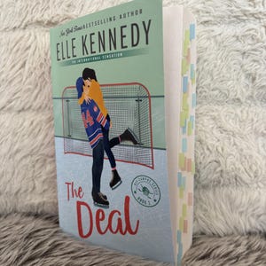 Puede incluir: Un libro de bolsillo titulado "The Deal" de Elle Kennedy. La portada presenta a una pareja abrazándose en una pista de hielo, con una portería de hockey de fondo. El libro tiene notas adhesivas de colores.