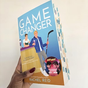 May include: A paperback book titled "GAME CHANGER" with a blue and orange cover. The cover features illustrations of two men, one holding a hockey stick. The book has colorful page markers and the author's name, Rachel Reid, is visible.