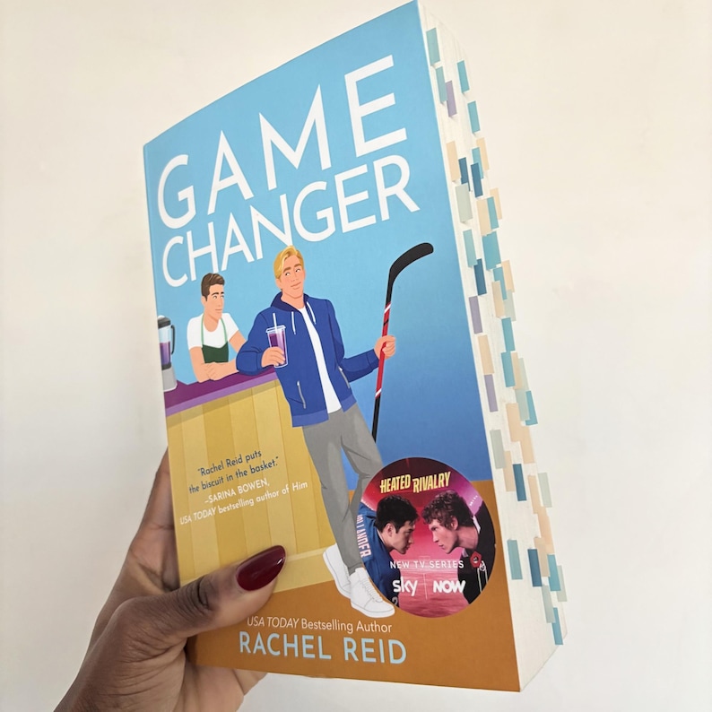 May include: A paperback book titled "GAME CHANGER" with a blue and orange cover. The cover features illustrations of two men, one holding a hockey stick. The book has colorful page markers and the author's name, Rachel Reid, is visible.