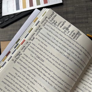 Puede incluir: Libro abierto con notas manuscritas y pesta&ntilde;as de colores. El texto incluye frases como "gonna have more details about the world" y "Can I have everyone's attention?" El libro est&aacute; sobre un escritorio con un teclado y pegatinas de anotaci&oacute;n.