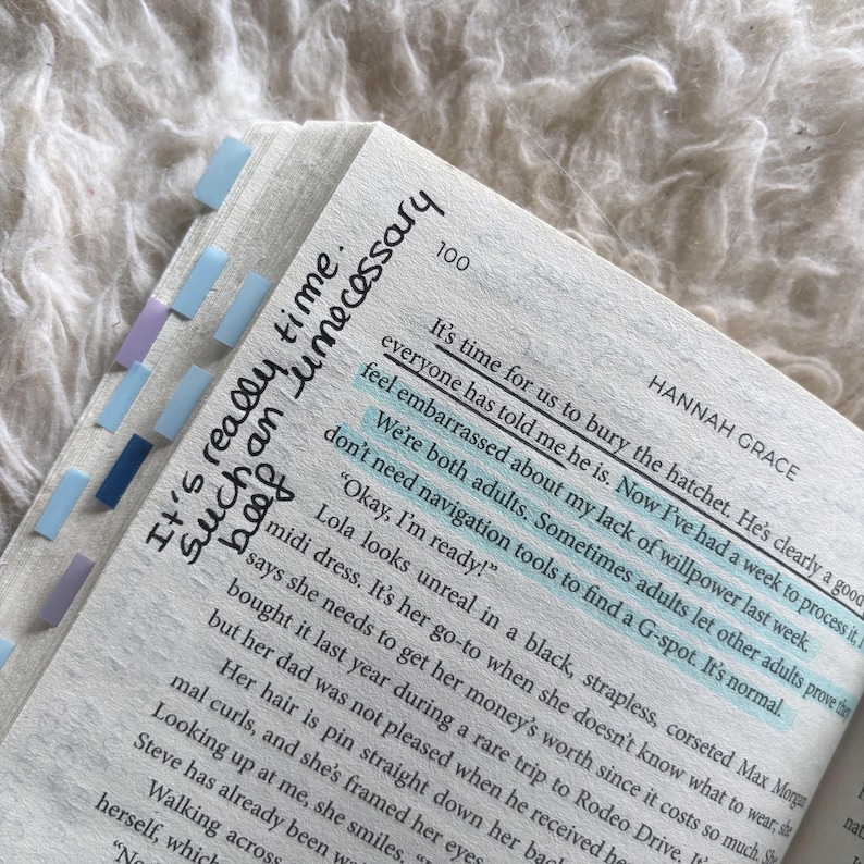 May include: A book page by Hannah Grace with page number 100, featuring printed text and handwritten notes in black ink. Several light blue, dark blue, and purple adhesive page markers are attached to the left edge of the paper.