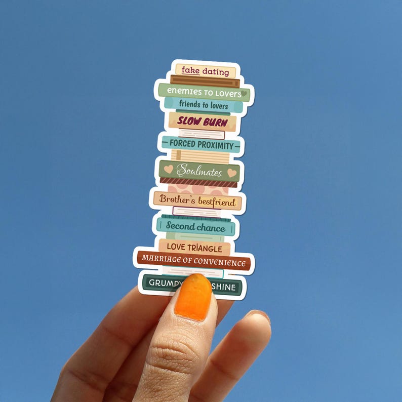 May include: A stack of colorful stickers with book-like designs, each labeled with romance novel tropes such as "fake dating," "enemies to lovers," and "slow burn." The stickers are held against a blue sky background.