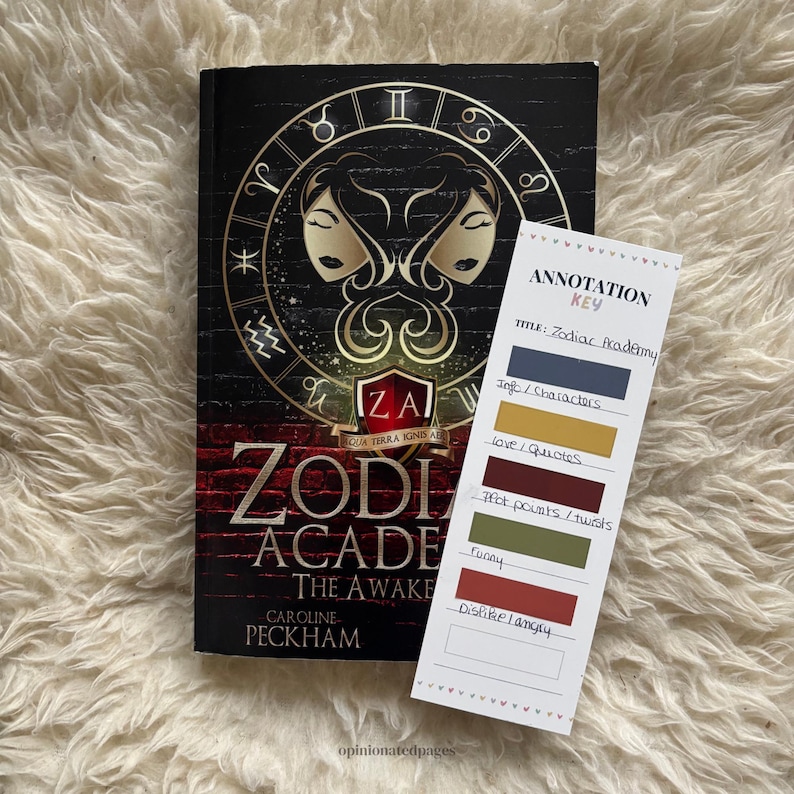 Puede incluir: Un libro de tapa dura negro titulado "Zodiac Academy: The Awakening" de Caroline Peckham, con s&iacute;mbolos dorados del zodiaco y un emblema central. Una clave de anotaci&oacute;n enumera categor&iacute;as como "Info/Personajes" y "Puntos de la trama/giros".