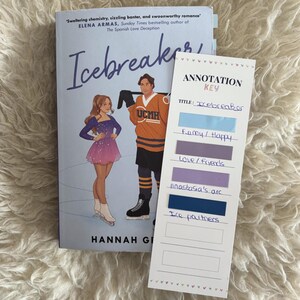 May include: The book Icebreaker by Hannah Grace features a cover illustration of a figure skater and a hockey player. Beside it is a white annotation key bookmark with colorful adhesive tabs labeled with handwritten notes for reading organization.