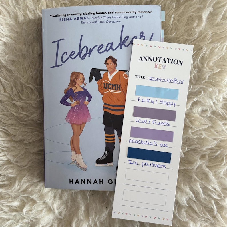 May include: The book Icebreaker by Hannah Grace features a cover illustration of a figure skater and a hockey player. Beside it is a white annotation key bookmark with colorful adhesive tabs labeled with handwritten notes for reading organization.