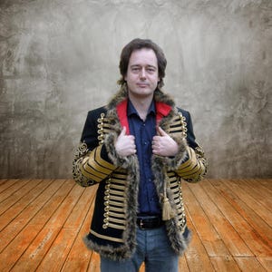 Op de afbeelding: Een zwarte jas met gouden afwerking en een kraag van imitatiebont. De jas heeft gouden versieringen op de mouwen en voorkant, met een rode kraag en een kwast. De persoon draagt een blauw overhemd en een spijkerbroek.