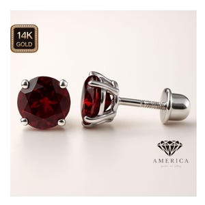 May include: A pair of stud earrings featuring round, deep red gemstones set in a silver metal. The earrings have a screw-back closure. The image also includes a gold-colored square with the text "14K GOLD" and a diamond logo with the word "AMERICA".