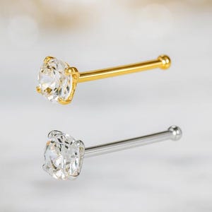 Könnte beinhalten: Zwei Nasenstecker, einer goldfarben und einer silberfarben, jeweils mit einem klaren, rund geschliffenen Edelstein. Der goldfarbene Stecker hat einen gelbgoldenen Stab und eine Fassung, der silberfarbene Stecker hat einen silberfarbenen Stab und eine Fassung.