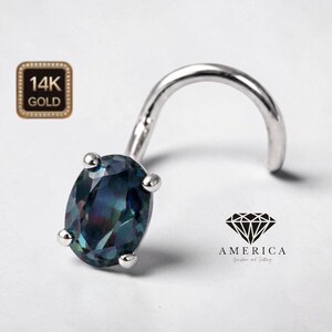 May include: A nose ring with a curved post and an oval-shaped, iridescent gemstone. The setting is silver-toned, with small, round accents securing the gem. A gold-colored square with "14K GOLD" is in the upper left corner. The text "AMERICA Gemstone and Setting" is in the lower right.
