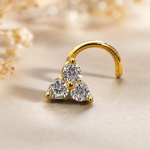 May include: A gold nose stud featuring a curved post and a triangular cluster of three clear, round-cut stones. The setting holds the stones in a tight formation, creating a bright, reflective surface that catches the light on the metal prongs.