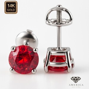 May include: A pair of stud earrings featuring a vibrant red gemstone set in a silver-colored metal. The earrings have a classic design with a round-cut stone and a secure post backing. The image also includes a 14K gold stamp.