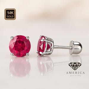 Puede incluir: Un par de pendientes de rubí con sello de oro de 14K. Los pendientes presentan una piedra preciosa roja oscura, redonda y facetada, engastada en un engaste de metal plateado. Un pendiente se muestra en posición vertical y el otro de lado.