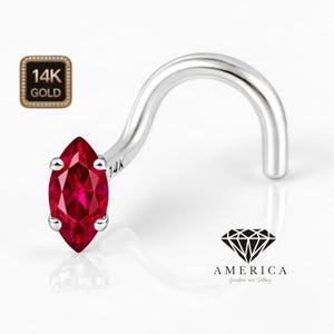 May include: A silver nose stud with a marquise-cut, deep red gemstone. The stud is curved and features a small, square gold emblem with the text "14K GOLD". The gemstone is held in place by four small prongs.