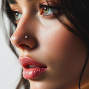May include: Close-up of a person's face, focusing on the nose and eye. A small, round, clear nose stud is visible on the nostril. The skin is smooth and the lips are a natural pink color. The eyes are green with long eyelashes.