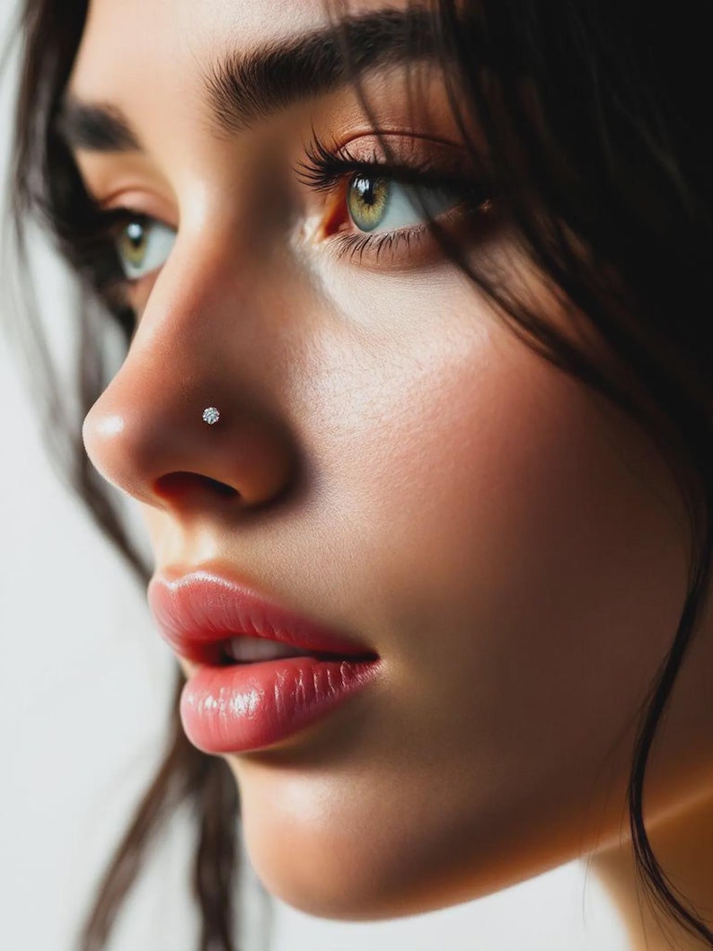 May include: Close-up of a person's face, focusing on the nose and eye. A small, round, clear nose stud is visible on the nostril. The skin is smooth and the lips are a natural pink color. The eyes are green with long eyelashes.