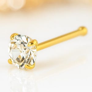 May include: A gold nose stud featuring a clear, round-cut gemstone. The gemstone is held in place by four gold prongs. The stud has a straight, slender gold post.