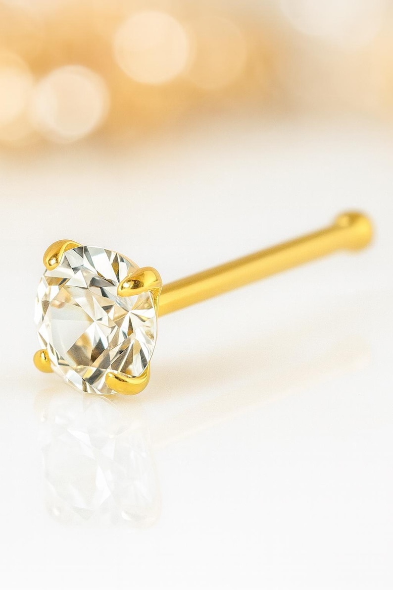 May include: A gold nose stud featuring a clear, round-cut gemstone. The gemstone is held in place by four gold prongs. The stud has a straight, slender gold post.