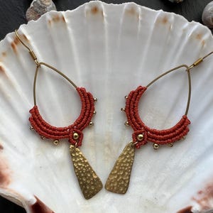 May include: A pair of teardrop-shaped earrings featuring rust-coloured macrame cord woven around a gold-toned wire frame. Each earring is accented with small gold-toned beads and a dangling, hammered gold-toned metal charm at the bottom centre.