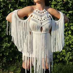 Puede incluir: Un top halter de macramé blanco con intrincados patrones de nudos, una abertura central en forma de lágrima en el pecho y largos flecos fluidos que se extienden desde los hombros hasta el dobladillo, creando una apariencia texturizada.