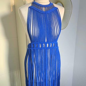 Könnte beinhalten: Ein leuchtend blaues Makramee-Halterkleid, das auf einer Schaufensterpuppe präsentiert wird. Das Kleid hat einen Neckholder-Ausschnitt, einen detaillierten Gürtel und lange Fransen. Das Kleid ist handgefertigt und scheint im Bohemian-Stil zu sein.