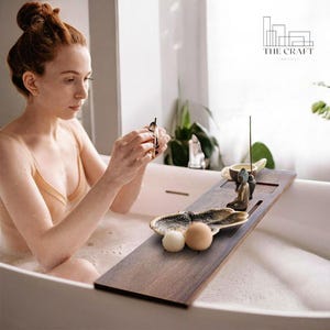 Puede incluir: Una bandeja de baño de madera con un plato decorativo en forma de concha, dos bombas de baño, un pequeño quemador de incienso y una pequeña estatua de Buda. La bandeja está sentada en el borde de una bañera blanca.