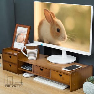 Puede incluir: Un organizador de escritorio de madera con cajones y un estante. El organizador es marrón y tiene recortes en forma de corazón en los cajones. Hay un monitor de computadora, una taza, un teléfono y un teclado en el escritorio.