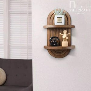 May include: A wooden wall shelf with a rounded arch design. The shelf has two tiers and is decorated with a small potted plant, a vintage phone, and a wooden figurine. The shelf is mounted on a white wall.