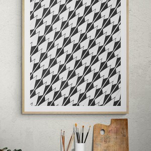 May include: A framed black and white geometric art print. The artwork features a repeating pattern of black and white arrow-like shapes. The print is displayed above a wooden table with art supplies.