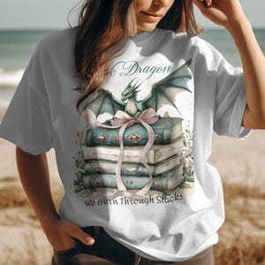 May include: White t-shirt with a blue dragon illustration sitting on a stack of books. The text "Book Dragon" and "We Burn Through Stacks" is printed on the shirt.