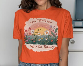 Puede incluir: Camiseta naranja con un gráfico de una puesta de sol y flores. El texto "Grow through what you go through" está impreso en la camiseta.
