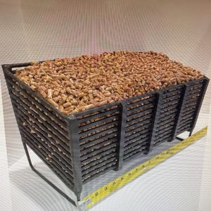 May include: A rectangular, dark metal bin filled with brown wood pellets. The bin has a slatted design and is supported by a metal frame. A yellow ruler is placed next to the bin, showing measurements in inches.