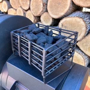 Op de afbeelding: Een zwarte metalen houtskoolmand gevuld met houtskoolbriketten staat op een zwarte smoker. De achtergrond toont een stapel gezaagde boomstammen, wat duidt op buiten grillen en barbecueën.