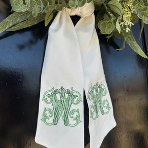 Peut inclure: Écharpe blanche avec monogramme "W" brodé en vert. Le foulard est fait d'un tissu doux et présente un motif floral décoratif autour de l'initiale. Le foulard est suspendu sur un fond sombre, avec de la verdure en arrière-plan.