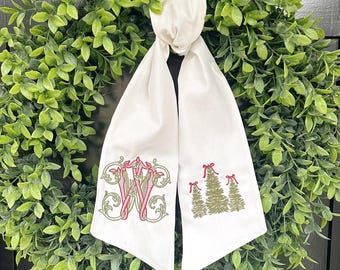 May include: A white fabric Christmas wreath sash with embroidered details. The sash features a large initial "W" in red and green, and three Christmas trees with red bows. The sash is tied to a green wreath.
