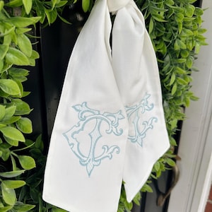 May include: White fabric door hanger with light blue embroidered monogram. The fabric is tied in a knot and hangs against a green wreath. The monogram is an ornate design, adding a touch of elegance to the door decor.