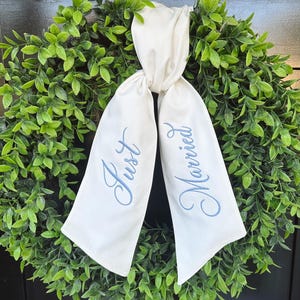 Peut inclure: Une couronne verte avec un ruban blanc noué en haut. Le ruban porte les mots "Just Married" brodés en écriture bleu clair. La couronne est faite de feuilles vertes.