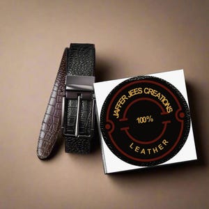 Op de afbeelding: Twee zwarte leren riemen met een zilveren gesp. De riemen liggen op een bruin oppervlak naast een witte doos met een zwarte en rode cirkel waarop "Jaffar Jees Creations 100% Leather" staat.