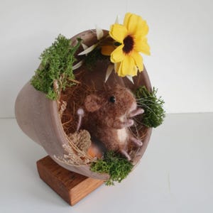 May include: A handcrafted felted mouse sculpture nestled in a tilted brown ceramic pot, adorned with green moss, dried plants, and a vibrant yellow flower. The sculpture rests on a small wooden base, creating a whimsical miniature scene.