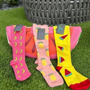 May include: Three pairs of knee-high socks with fruit patterns. One pair is pink with yellow bananas, another is pink with yellow pineapples, and the third is yellow with red watermelon slices. Each pair has a tag that says "SCRUB LIFE".