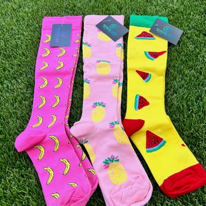 May include: Three pairs of knee-high socks with fruit patterns. One pair is pink with yellow bananas, another is pink with yellow pineapples, and the third is yellow with red watermelon slices. Each pair has a tag that says "SCRUB LIFE".