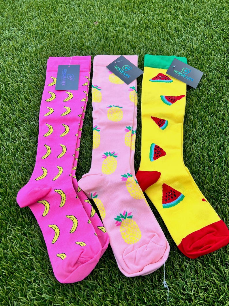 May include: Three pairs of knee-high socks with fruit patterns. One pair is pink with yellow bananas, another is pink with yellow pineapples, and the third is yellow with red watermelon slices. Each pair has a tag that says "SCRUB LIFE".