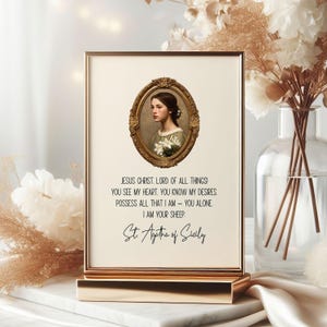 May include: A framed print with a gold-colored frame and stand. The print features an oval portrait of a woman in a gold frame, with text below that reads "JESUS CHRIST. LORD OF ALL THINGS!" and "St. Agatha of Sicily."