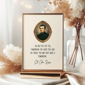 May include: A framed print with a gold-colored frame and stand. The print features a portrait in an oval frame above the quote: "DO NOT PUT OFF TILL TOMORROW THE GOOD YOU CAN DO TODAY. YOU MAY NOT HAVE A TOMORROW." Signed "St. John Bosco."