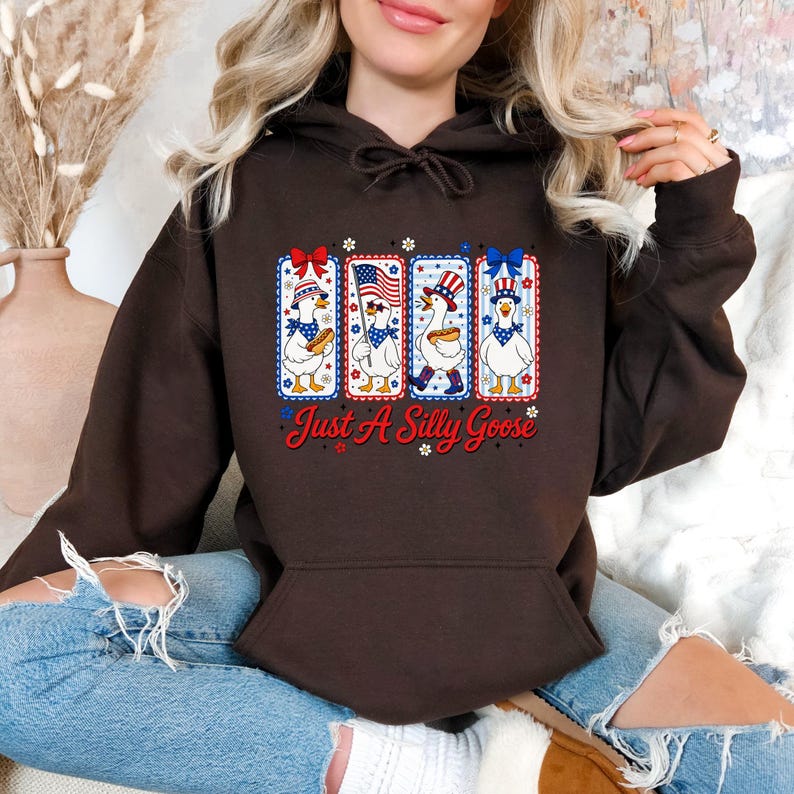 A woman wearing a brown hoodie with a graphic design of four cartoon geese and the text "Just A Silly Goose" printed on it.
