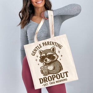 A woman is holding a white tote bag with a graphic design featuring a raccoon wearing sunglasses and holding a drink.
