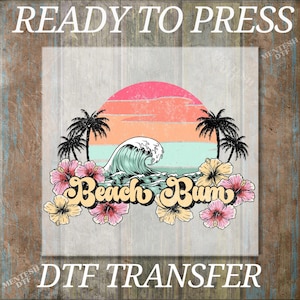 May include: A digital transfer design featuring a sunset over a beach with a wave crashing. The text "Beach Bum" is in a retro font with hibiscus flowers surrounding it.