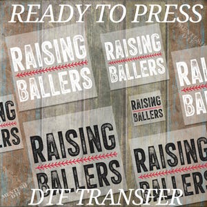 May include: Five sheets of iron-on transfer paper with the text "Raising Ballers" in white and black lettering with a red baseball stitch design. The text "Ready to Press" and "DTF Transfer" are also visible.