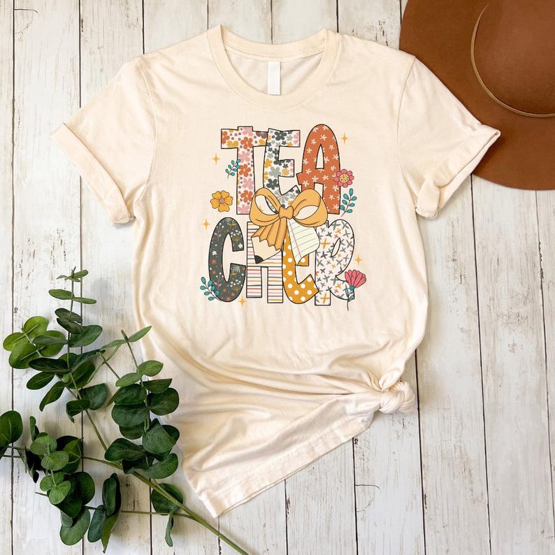 A beige t-shirt with a colorful graphic design featuring the text "TEACHER" in a playful font, surrounded by various decorative elements such as flowers and leaves.
