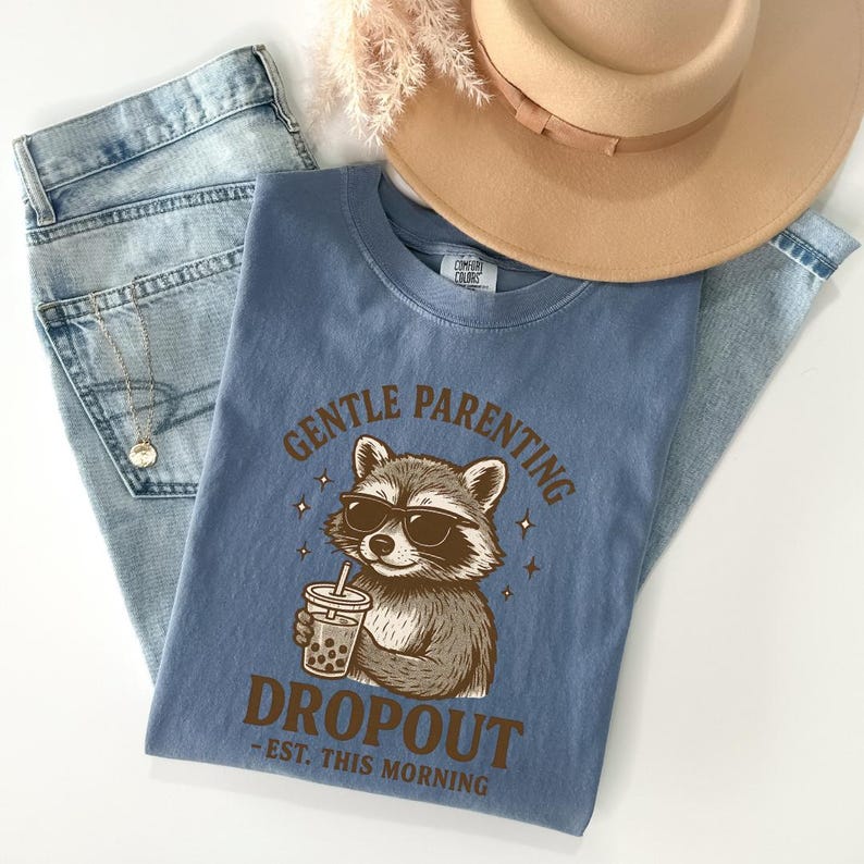 A blue t-shirt with a raccoon graphic and text, along with a beige hat and denim jeans, are laid out on a white surface.